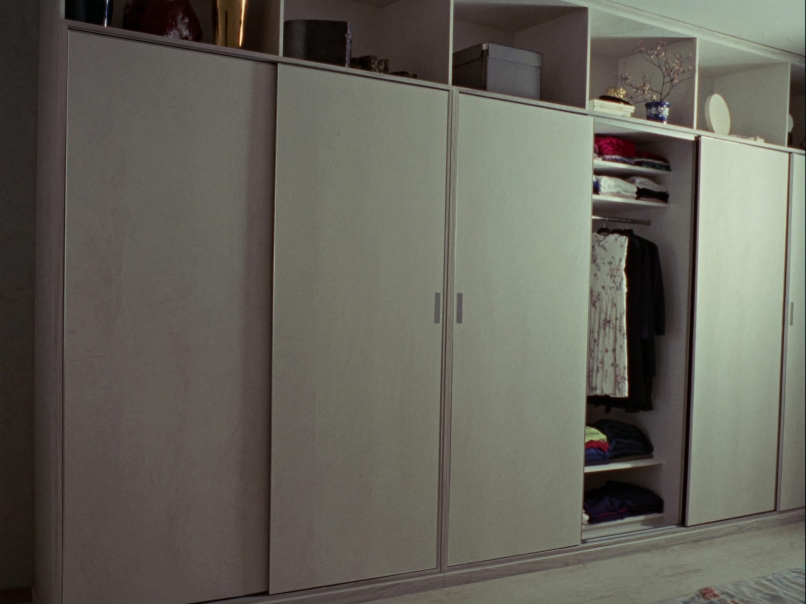 A built-in bedroom wardrobe with full height sliding doors in a light grey woodgrain melamine finish, shelving and a hanging rail visible through an open door. Illustrative concept visual, not a real photograph.