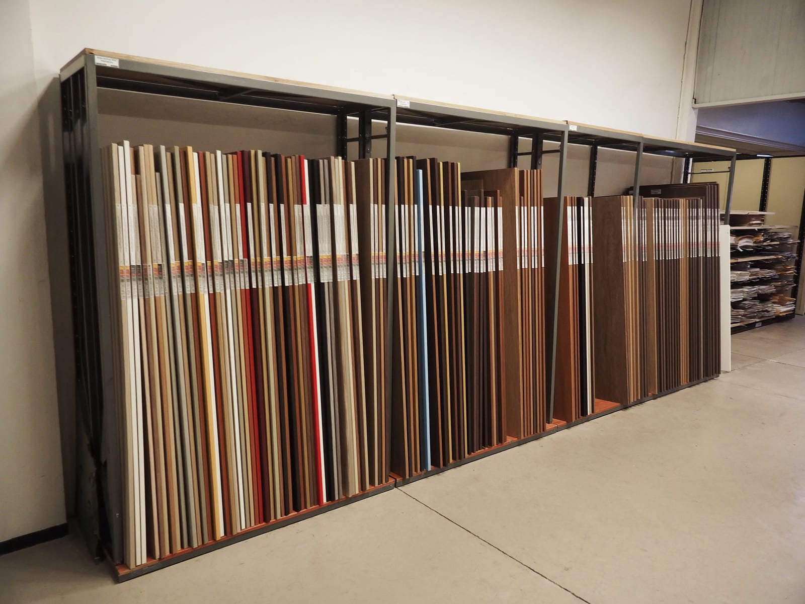 A tall steel rack in a board supplier warehouse holding full size melamine faced sheets standing on edge, their coloured and woodgrain faces showing in a row. Illustrative concept visual, not a real photograph.
