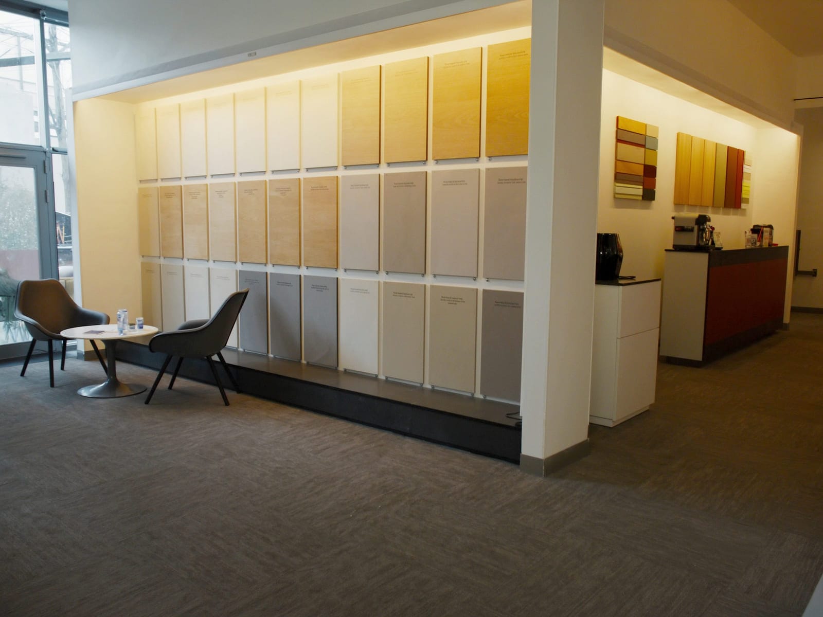 A corner of a modern board showroom with two chairs and a round table beside a large sample display wall, and a coffee machine on a side counter. Illustrative concept visual, not a real photograph.