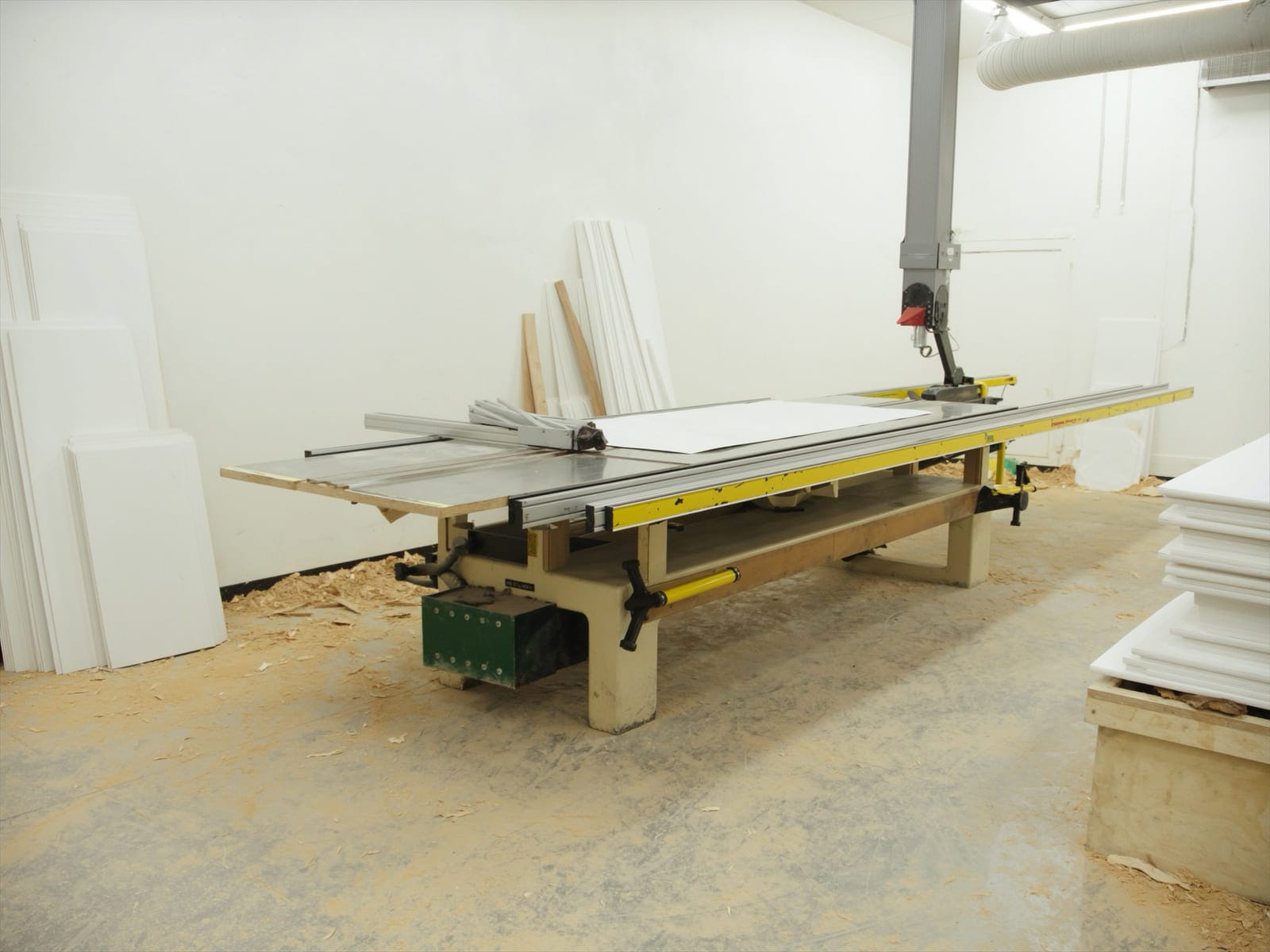 An industrial beam panel saw in a board supplier workshop with a stack of freshly cut white melamine panels on its table and offcuts stacked to one side. Illustrative concept visual, not a real photograph.
