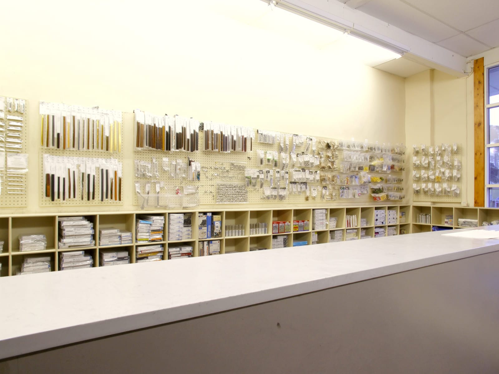The service counter of a hardware and board supply store, with shelves and pegboard walls behind it stocked with cabinet handles, hinges and fittings in bins. Illustrative concept visual, not a real photograph.