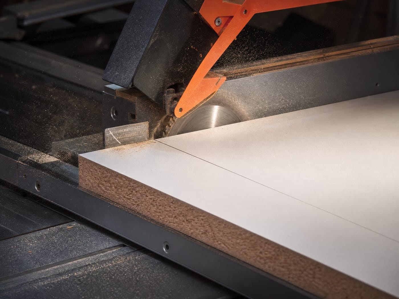 A panel saw blade cutting a white melamine-faced board to size, sawdust spraying from the cut.